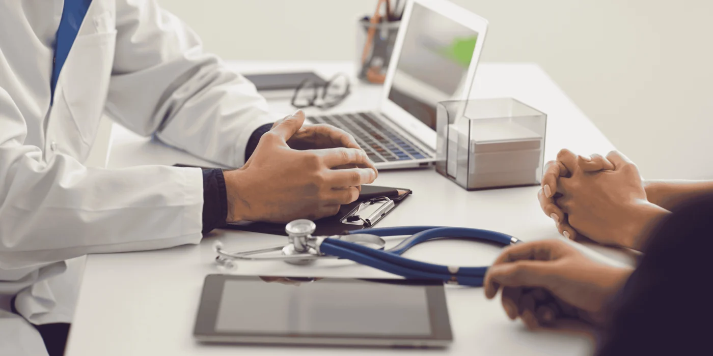 A doctor in a white lab coat consulting with a patient at a desk. The doctor’s hands are visible as they hold a clipboard, while a stethoscope and tablet lie on the desk. The patient’s hands are clasped together across the table, with a laptop in the background.